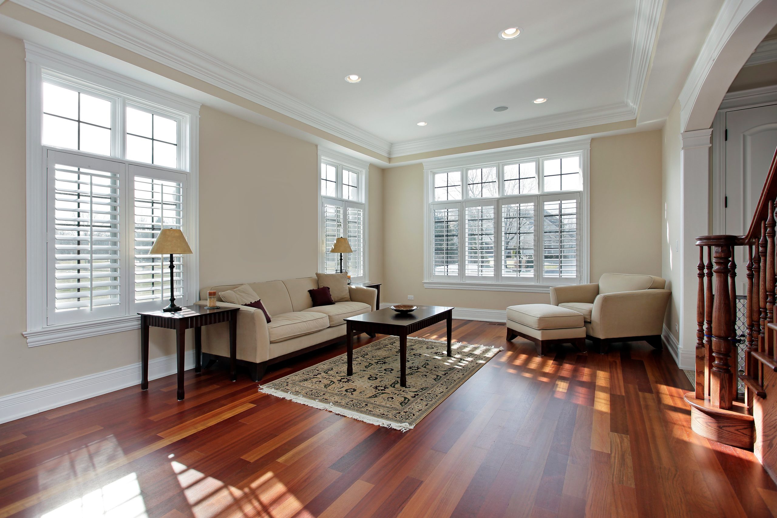 Sunlit living room with cream sofas and dark wooden flooring; rich reddish-brown planks anchor the neutral space beneath large shuttered windows